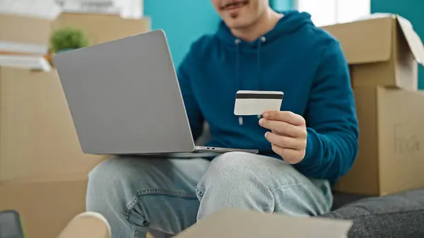 Young hispanic man shopping with laptop and credit card sitting on sofa at new home