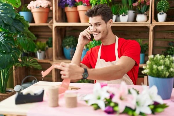 Young hispanic man florist talking on smartphone holding gift lace at flower shop
