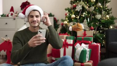 Yılbaşı hediyeleri, kulaklıkları ve akıllı telefonları olan yakışıklı genç adam Noel 'i evde kutluyor.