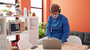 Kulaklıklı bir adam evde dizüstü bilgisayarla çalışırken telefonla konuşuyor.