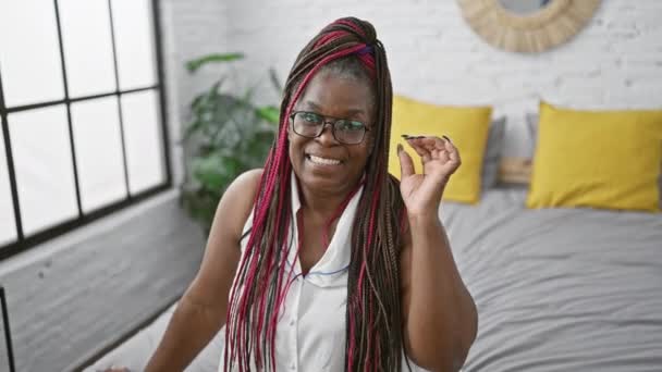彼女の快適なベッドに座っている陽気なアフリカ系アメリカ人女性は 自信を持って彼女のホームレスの寝室で幸せな笑顔をスポーツしながらメガネを外します — ストック動画