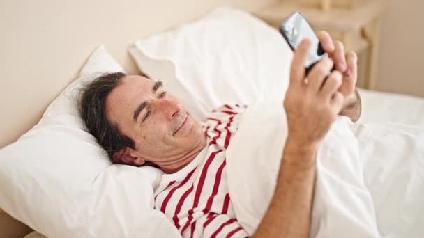 Mann Mittleren Alters Mit Smartphone Auf Bett Liegend Lächelt Ins — Stockvideo