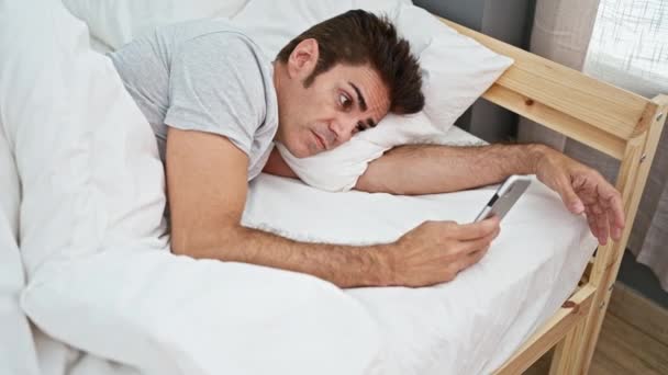 寝室でスマートフォンを使用して悲しい表情でベッドに横たわる若いヒスパニックの男 — ストック動画