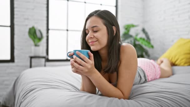 自信と美しい若いヒスパニックの女性はベッドで快適に横たわり 彼女の居心地の良い寝室でコーヒーの新鮮なカップの活気に満ちた朝の香りを楽しみます — ストック動画