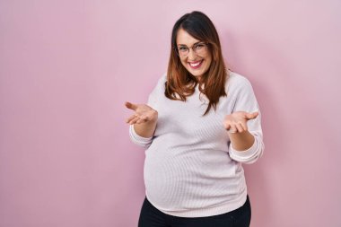 Pembe arka planda durup gülümseyen, yardım eli uzatan ve kabullenen hamile bir kadın.. 