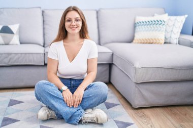 Oturma odasında, yüzünde mutlu ve havalı bir gülümsemeyle yerde oturan beyaz bir kadın. şanslı kişi. 