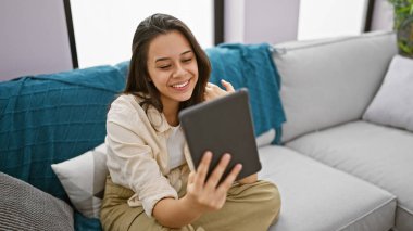 Karşı konulmaz bir şekilde kendine güveni olan genç İspanyol bir kadın rahat kanepesinde dinlenirken mutlu bir şekilde dokunmatik ped kullanıyor. Oturma odasının rahat ortamında pozitiflik yayıyor..