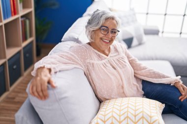 Orta yaşlı, kır saçlı, gülümseyen bir kadın. Evdeki kanepede oturuyor.