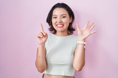 İspanyol genç bir kadın pembe arka planda duruyor. Altıncı parmaklarını gösterip işaret ederken kendinden emin ve mutlu bir şekilde gülümsüyor.. 