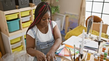 Sanat stüdyosunun içinde, örgü örgülü güzel Afrikalı Amerikalı kadın ressam resim hobisine dalmışken dizüstü bilgisayarından rengini seçiyor.