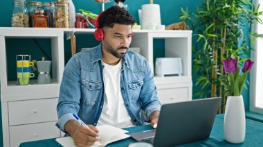 Afro-Amerikalı adam yemek odasında dizüstü bilgisayar ve kulaklık kullanıyor.