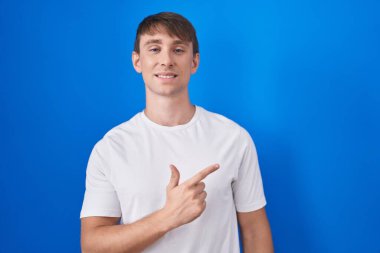 Kafkasyalı sarışın adam mavi arka planda neşeli, yüzünde bir gülümsemeyle mutlu ve doğal bir ifadeyle elini ve parmağını yana kaldırdı. 