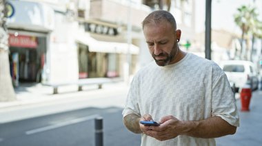 Ciddi görünümlü, çekici bir genç adam akıllı telefonuna dalmış güneşli bir şehir sokağında dikiliyor, serin ve konsantre bir yaşam tarzı sergiliyor.