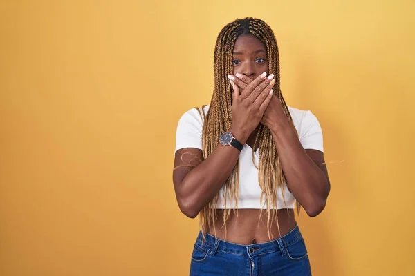 黄色い背景の上に立っている編組の髪を持つアフリカ系アメリカ人女性は 間違いのために手で口を覆うショックを受けました シークレットコンセプト — ストック写真