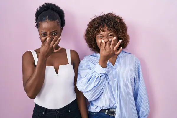 ピンクの背景の上に立っている2人のアフリカ人女性は 臭いと嫌な臭いを嗅ぎ 鼻に指で息を止めます 悪い匂いがする — ストック写真