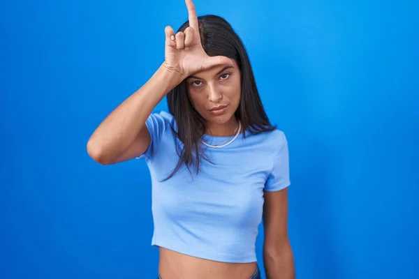ブルネット 青い背景の上に立っている若い女性は 失われたジェスチャーを嘲笑し侮辱している額に指を持つ人々の楽しみを作ります — ストック写真