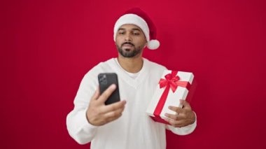 Noel Baba şapkalı mutlu melez adam elinde akıllı telefon ve kırmızı arka planda dururken hediyeyle selfie çekiyor.