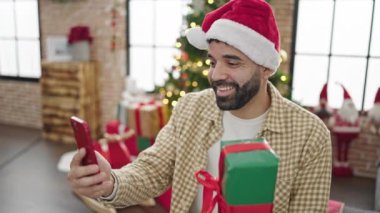 Kırmızı Noel Baba şapkalı adam hediyesini tutuyor ve Noel 'de cep telefonuyla selfie çekiyor.