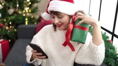 Kafkasyalı genç bir kadın akıllı telefon kullanıyor. Noel ağacının yanında oturuyor.