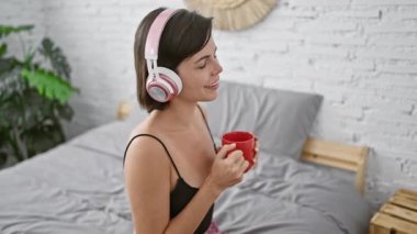 Sabah saadeti, kendine güvenen, güzel, genç İspanyol kadın espresso yudumluyor, yatakta müzik dinliyor, rahat pijama ve battaniyeye sarılmış, gülümsüyor, evinde rahat bir şekilde dinleniyor.