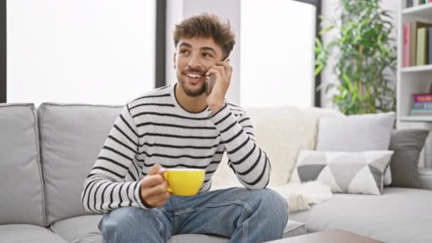 ハッピーアラビアの男は リラックスした朝食を楽しんで 座って コーヒーカップを手に 彼の居心地の良い家で スマートフォンで自信を持って話す笑顔 — ストック動画