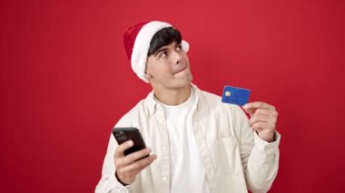 Akıllı telefon ve kredi kartıyla alışveriş yapan genç İspanyol adam izole edilmiş kırmızı arka planda noel şapkası takıyor.