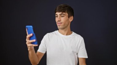 Neşeli, genç, İspanyol asıllı bir adam ayakta dururken, telefonda konuşurken ve video görüşmesinde izole edilmiş, havalı bir siyah duvar arka planına karşı eğlenirken kendine güvenerek gülümsüyor.