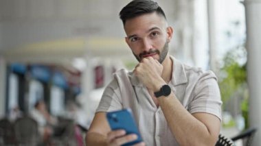 Genç İspanyol adam akıllı telefon kullanıyor. Kahve dükkanının terasında oturmuş gülümsüyor.