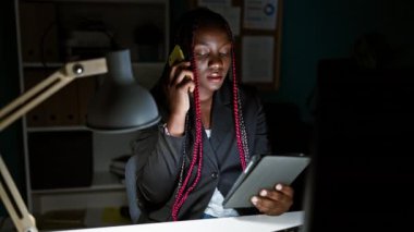 Konsantre olmuş Afro-Amerikan kadın patron akıllı telefondan iş konuşuyor, ofisin iç kısımlarında dokunmatik ped üzerinde çalışıyor.