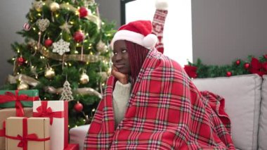 Saçları örülmüş Afrikalı bir kadın Noel ağacının yanında, koltukta oturmuş üzgün üzgün eve bakıyor.