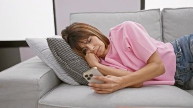 Çevrimiçi dünyada kaybolmuş, evdeki oturma odasındaki kanepede rahatça uzanan parlak, genç İspanyol bir kadın. Daktilo yazma, ciddi ifade, kısa saç bir yetişkin kenarı ekleme.