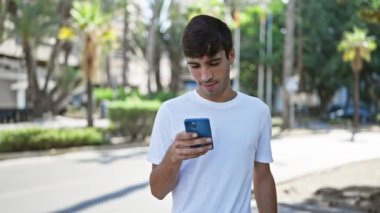 Neşeli İspanyol asıllı genç adam şehir parkında gezici bir galibiyeti coşkuyla kutluyor, doğanın yeşili arasında akıllı telefon teknolojisini kullanarak mutlu bir şekilde mesaj yazıyor.