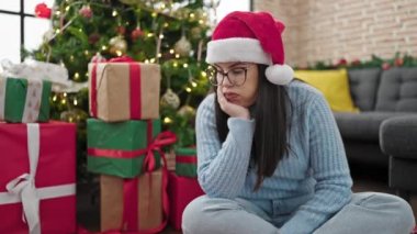 Genç İspanyol kadın Noel şapkası takarak yerde oturuyor. Evde sıkılmış görünüyor.