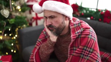 Genç kel adam Noel ağacının yanında oturmuş üzgün üzgün bakıyor.