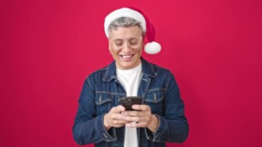 Kafkasyalı genç adam yılbaşı şapkası takıyor. Akıllı telefonu kullanarak izole edilmiş kırmızı arka planda kutlama yapıyor.
