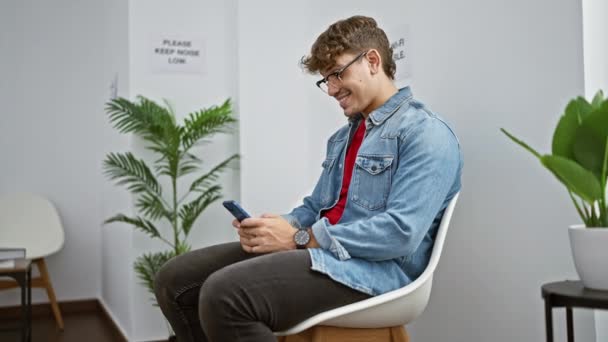 快乐的年轻人坐在候车室的椅子上 用智能手机在室内环境中传递着胜利的信息 — 图库视频影像