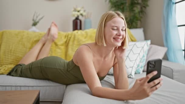 自宅のソファーに横たわっているスマートフォンでセルフィー写真を撮る若い金髪の女性 — ストック動画