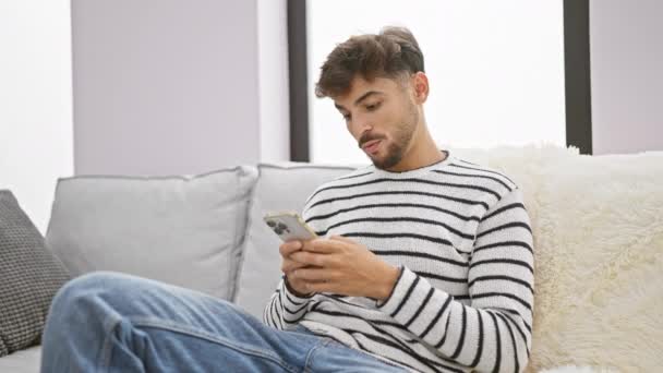 Překvapený Mladý Arabský Muž Ukazuje Smartphone Zatímco Radostně Sedí Pohovce — Stock video