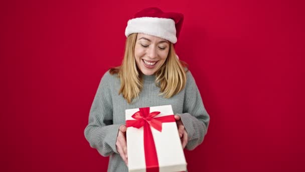 Młoda Blondynka Uśmiechnięta Pewnie Trzymając Prezent Świąteczny Nad Odizolowanym Czerwonym — Wideo stockowe