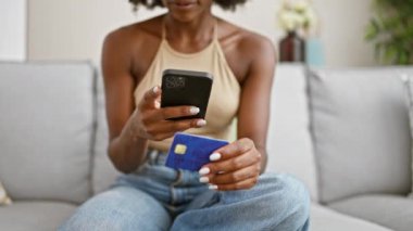 Afrikalı Amerikalı bir kadın evdeki kanepede oturan akıllı telefonuyla kredi kartına fotoğraf çekiyor.