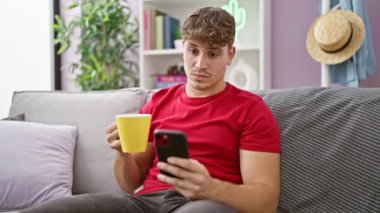 Şaşırmış genç İspanyol adam evde takılıyor, akıllı telefon kullanıyor, oturma odasındaki koltukta kahve içiyor.