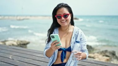 Genç Çinli kadın turist deniz kenarında elinde su şişesiyle akıllı telefondan konuşuyor.