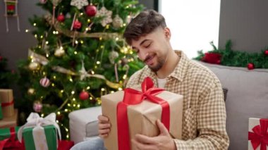 Genç Arap adam elinde hediyeyle evde Noel ağacının yanında oturuyor.