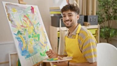 Canlı bir üniversite sanat stüdyosunda ustalıkla gülümseyen kendine güvenen genç Arap sanat öğrencisi, yaratıcılığını fırça ve paletle kullanıyor.