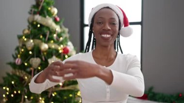 Afro-Amerikan kadın kalp hareketi yapıyor. Evde Noel ağacının yanında duruyor.