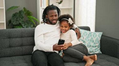 Afro-amerikan baba ve kızı akıllı telefon kullanıyor kanepede oturuyor ve gülümsüyor.