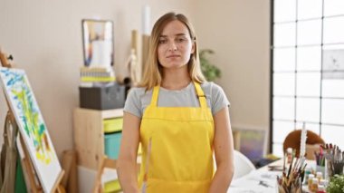 Kendine güvenen genç sarışın sanatçı, kollarını kavuşturup gülümsüyor, sanat stüdyosunun yaratıcı fırtınasının ortasında duruyor..