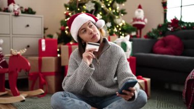 Akıllı telefon ve kredi kartı kullanan genç ve güzel bir İspanyol kadın evdeki Noel ağacının yanında oturuyor.