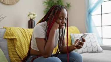 Mutsuz Afro-Amerikan kadın akıllı telefon kullanıyor, oturma odasındaki kanepede oturuyor, evdeki üzüntüsünü dile getiriyor.