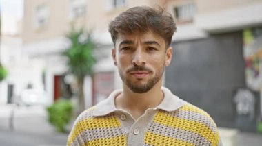 Kendine güvenen genç Arap adam, neşe ve pozitiflik saçıyor, bir şehir caddesinde duruyor, güneş gözlükleriyle göz kamaştırıcı bir şekilde yakışıklı, bulaşıcı gülüşü şehir geçmişini aydınlatıyor..